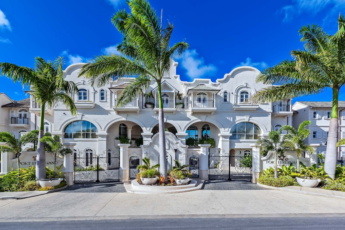 The exterior of a luxury villa is showcased, featuring elegant architectural details and multiple balconies. Lush palm trees frame the entrance, while a winding driveway leads to an ornate gated entry. The clear blue sky enhances the vibrant colors of the building.