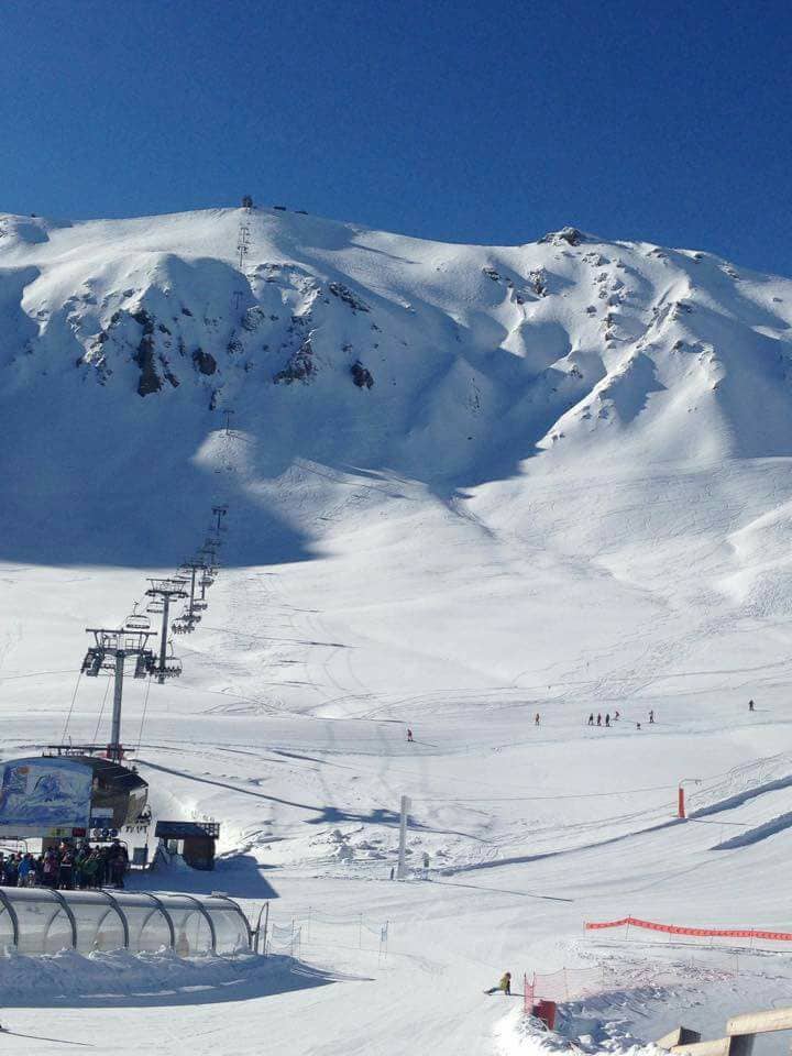 A snowy landscape is shown, featuring gentle slopes and ski lifts ascending the mountainside. The bright blue sky complements the white expanse of snow, creating a serene winter setting. A few skiers are visible in the distance, enjoying the slopes.