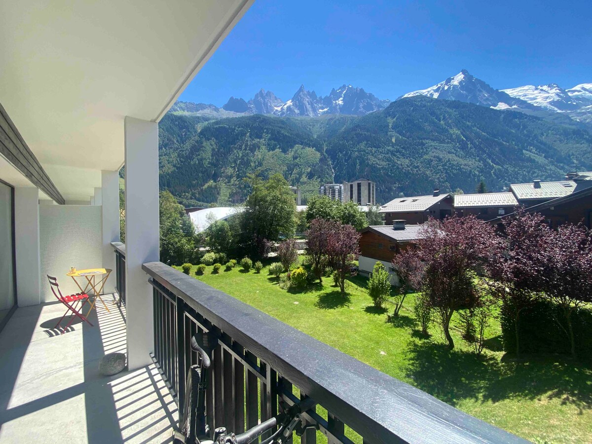 A large balcony features a table and chairs, with a backdrop of lush green gardens and mountainous terrain. Majestic peaks rise in the distance, covered in snow, under a clear blue sky. The space is ideal for outdoor relaxation and enjoying scenic views.