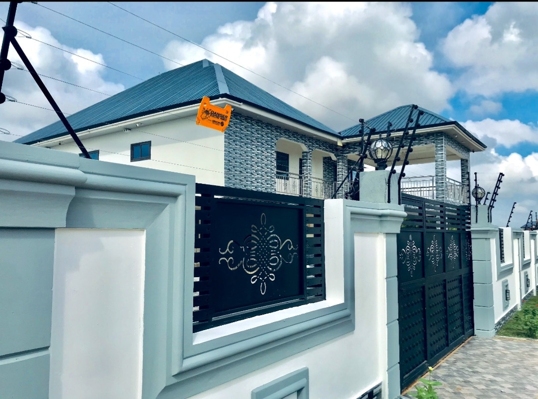 The exterior of the villa is presented, showcasing a modern design with a pitched roof and decorative wall elements. A gated entrance is visible, lined with wrought iron work and surrounded by landscaped grounds, while a vibrant sign indicates the property's name.