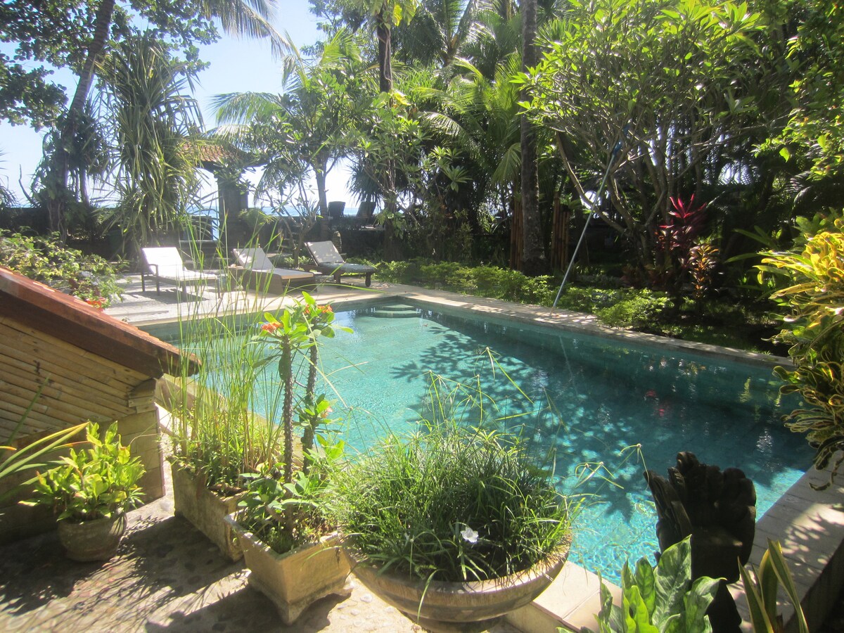 A serene pool is surrounded by lush tropical foliage, providing a sense of privacy. Sun loungers are positioned nearby, inviting relaxation. The clear blue water reflects the sunlight, creating a tranquil setting amidst the vibrant greenery.