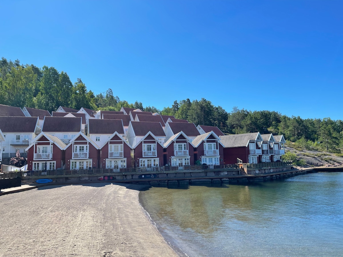 A row of charming red buildings is positioned along a sandy beach with clear water. Trees are visible in the background, offering greenery. Some buildings have balconies facing the water, providing a serene view of the sheltered bay.