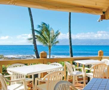 A dining area is visible with several white plastic tables and chairs arranged on a wooden deck. The ocean view showcases clear blue water and palm trees swaying gently in the breeze, creating a serene seaside atmosphere.