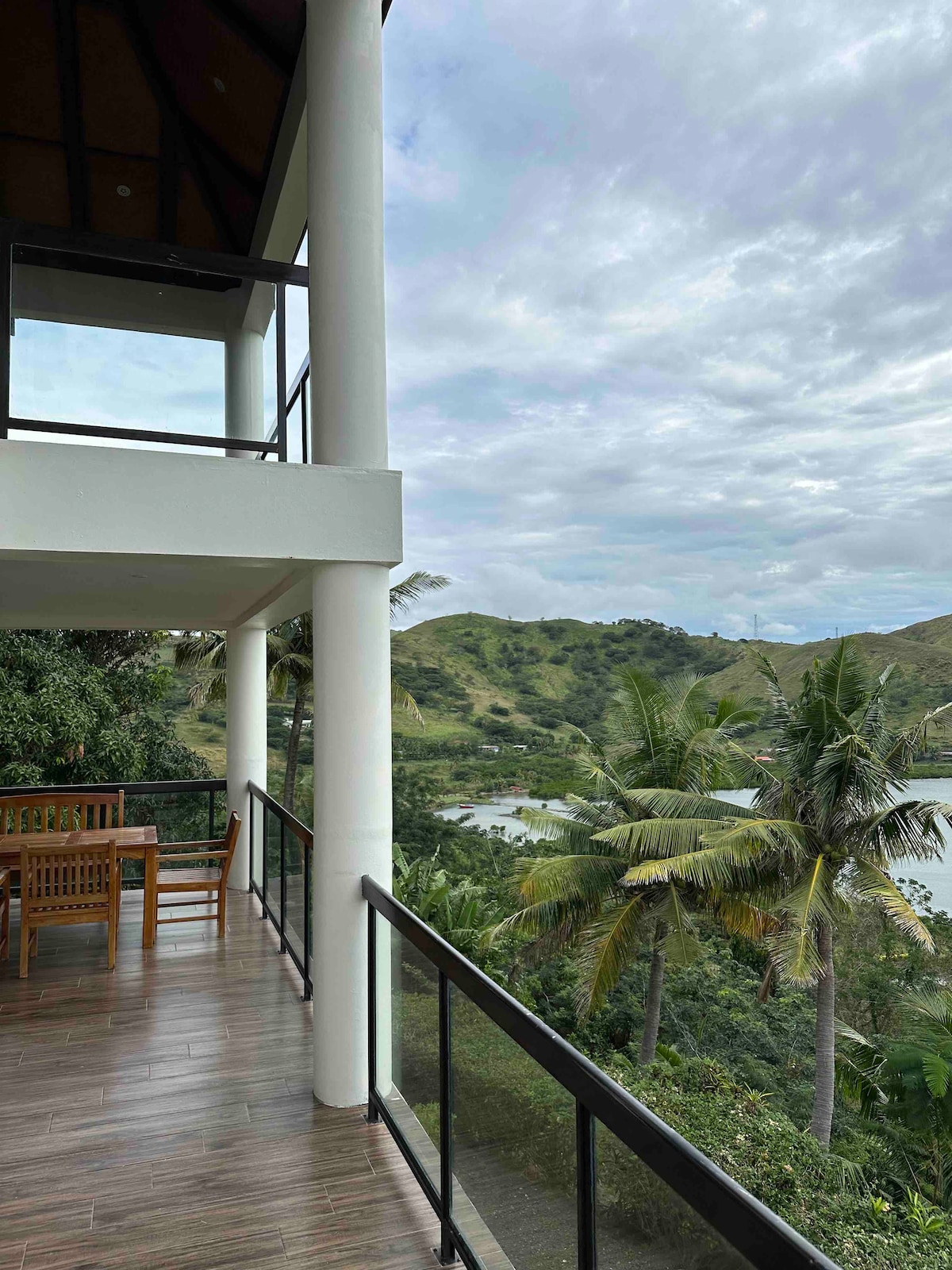 A spacious balcony is shown with a wooden dining table and chairs overlooking lush green hills and a winding river. The view features palm trees gently swaying in the breeze under a cloudy sky, providing a serene setting for relaxation.