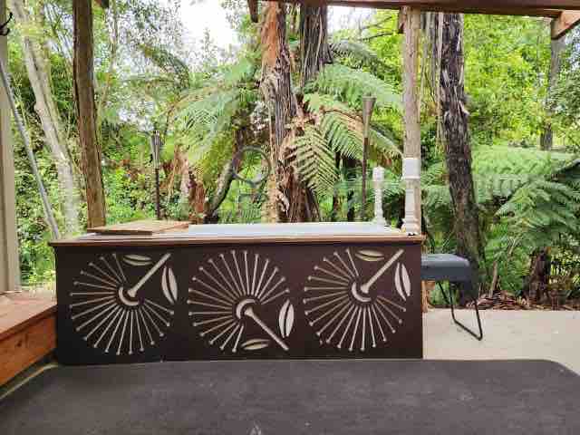 An outdoor bath is positioned on a spacious deck, complemented by artistic designs on its wooden structure. Lush greenery and native ferns create a serene backdrop, contributing to a tranquil setting surrounded by nature.