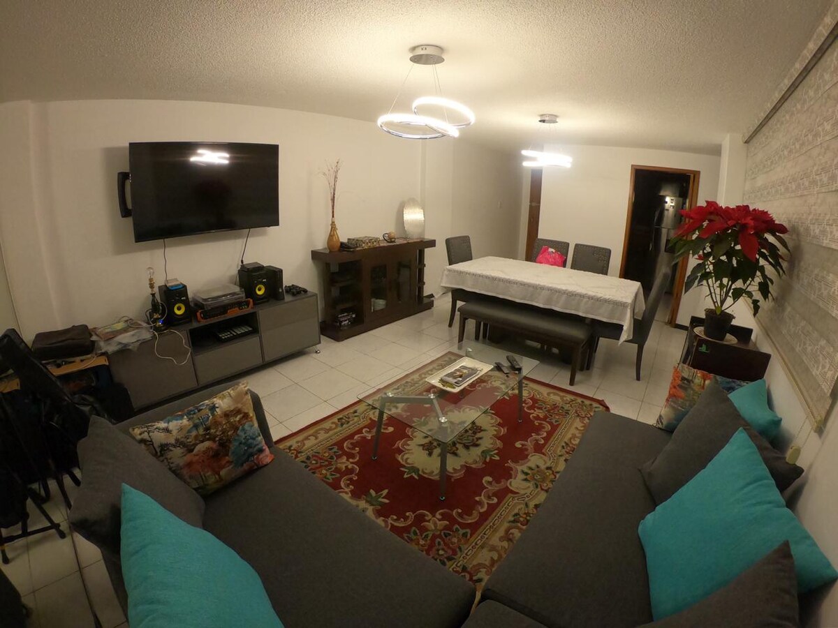 A spacious living area is featured with a medium-sized sofa adorned with bright cushions. A large television is mounted on the wall, alongside a decorative cabinet. A dining table accommodates multiple chairs, and a rug adds a touch of color beneath a glass coffee table.