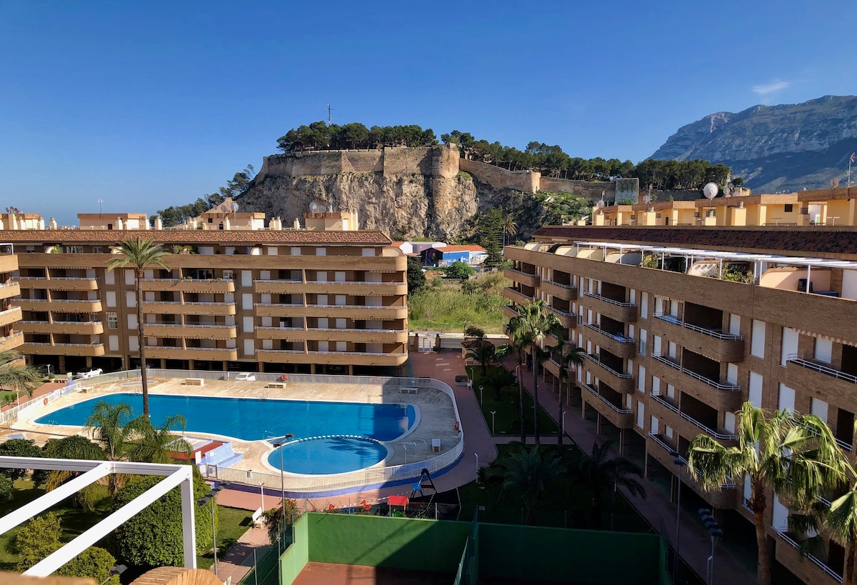 A spacious view of the apartment complex showcases the large swimming pool surrounded by palm trees. In the background, the historical castle and the mountain are visible, enhancing the scene. The architecture of the buildings features warm tones and multiple balconies.