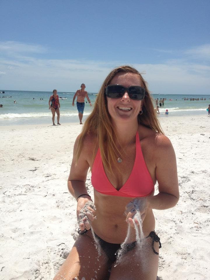 A sandy beach scene is observed with clear blue skies and gentle waves lapping at the shore. People are seen enjoying the water in the background, while an individual is seated in the foreground, playing with sand. The beach is lively, filled with sunbathers and beachgoers.