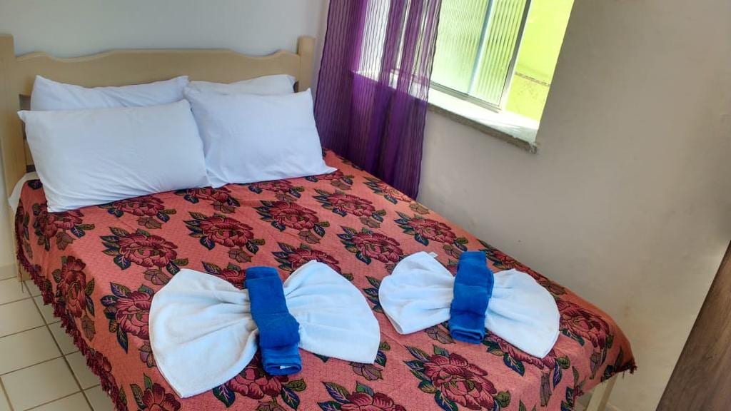 A cozy bedroom features a double bed adorned with a floral-patterned blanket. Two neatly arranged white towels are displayed at the foot of the bed, while natural light filters in through a partially opened window covered by sheer purple curtains.