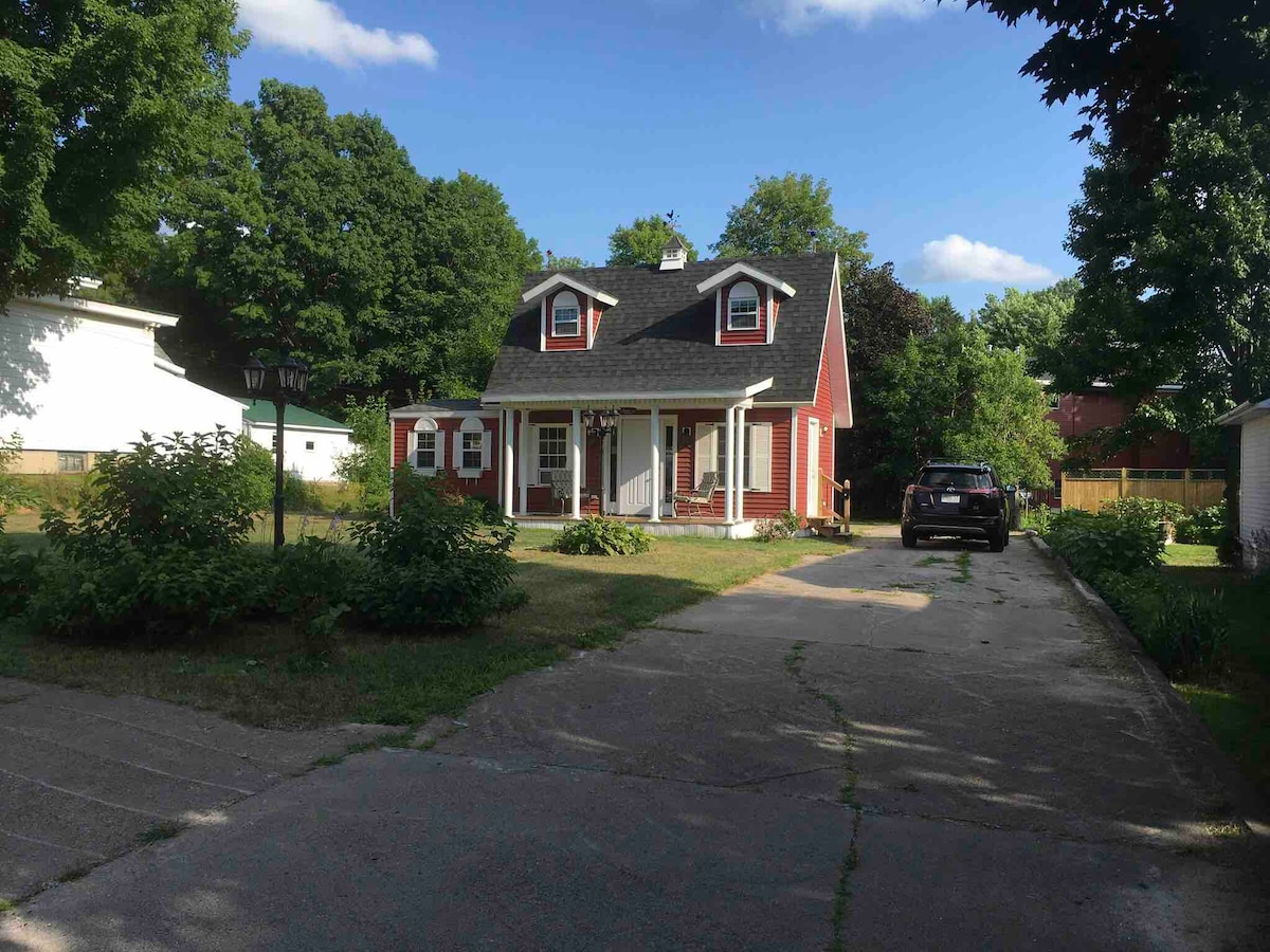 A charming two-story house features a red exterior and a welcoming front porch. The driveway, lined with greenery, leads to the entrance. Lush trees and bushes surround the property, providing a natural setting in a peaceful residential area.