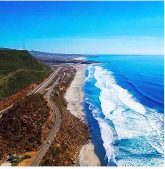 A sweeping aerial view captures the coastline, showcasing a sandy beach bordered by vibrant blue ocean waves. Rolling hills frame the scene, with a road meandering along the shore, inviting exploration of the picturesque landscape.