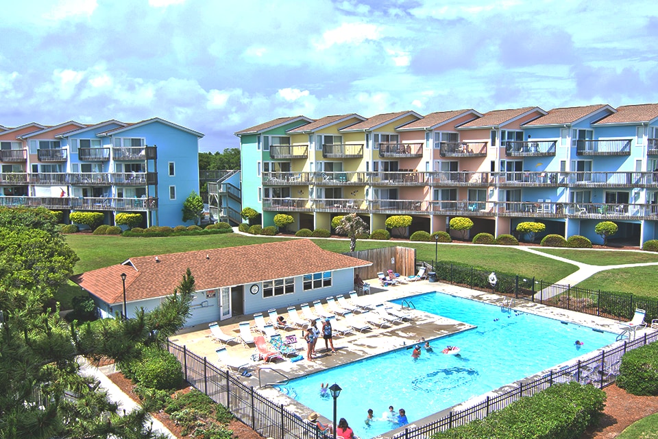 A vibrant pool area is surrounded by well-maintained grounds, with colorful, two-story condominium buildings in the background. Sun loungers are arranged around the pool, while guests enjoy recreational activities within the water. Lush greenery adds to the inviting ambiance of the complex.