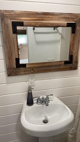 A compact sink is positioned below a wooden-framed mirror, reflecting the bright bathroom space. A soap dispenser is placed beside the faucet, and a towel bar is mounted on the wall above. Light streams through a nearby window, enhancing the clean, airy atmosphere.