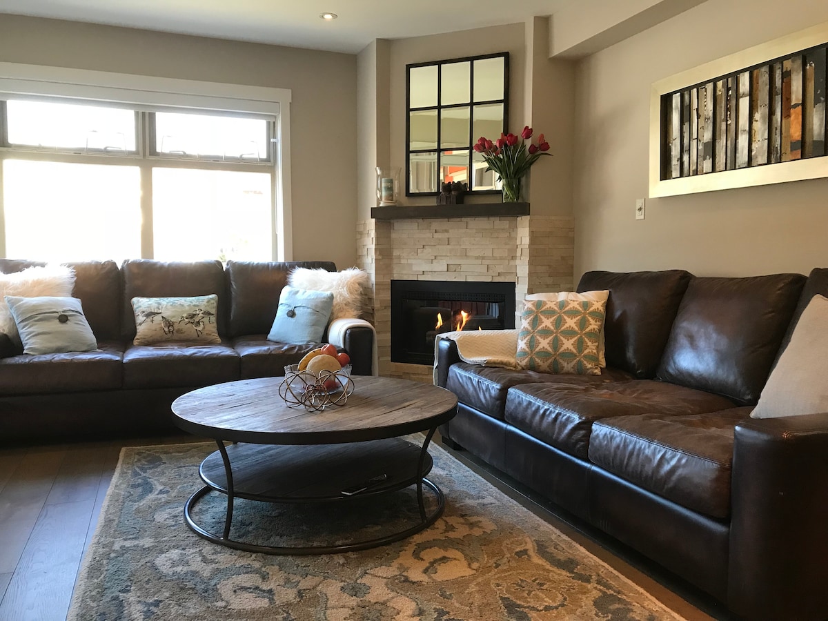 A cozy living room features two large leather sofas arranged around a circular wooden coffee table. A soft area rug lies beneath. A gas fireplace is situated against a stone wall, with natural light streaming through large windows adorned with decorative pillows and a vase of flowers.
