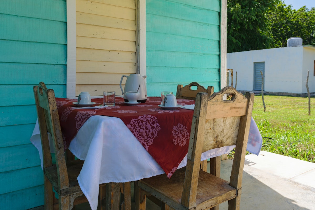 A wooden table is set outside under a colorful blue and white building. The table features a red tablecloth with white patterns, accompanied by several chairs. A teapot and cups are arranged on the table, inviting guests to enjoy a meal or drink in a serene outdoor setting.