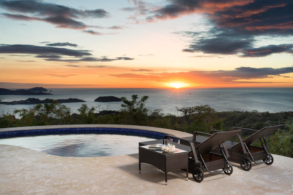 A tranquil pool area is pictured, with three lounge chairs positioned by the edge. A coffee table holds refreshments, set against a backdrop of a vibrant sunset over the ocean and distant hills, creating a serene atmosphere.