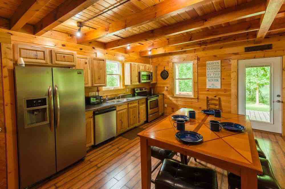 The fully equipped kitchen features wooden cabinetry with modern appliances, including a stainless steel refrigerator and oven. A dining table is set with blue plates, and natural light filters through the windows, illuminating the rustic wood beams and warm tones of the space.