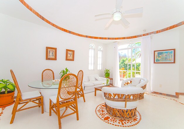 A bright and airy living area is surrounded by large windows allowing natural light to fill the space. A round glass dining table is set with white chairs, while two stylish wicker chairs invite relaxation. Plants add a touch of greenery to the room.