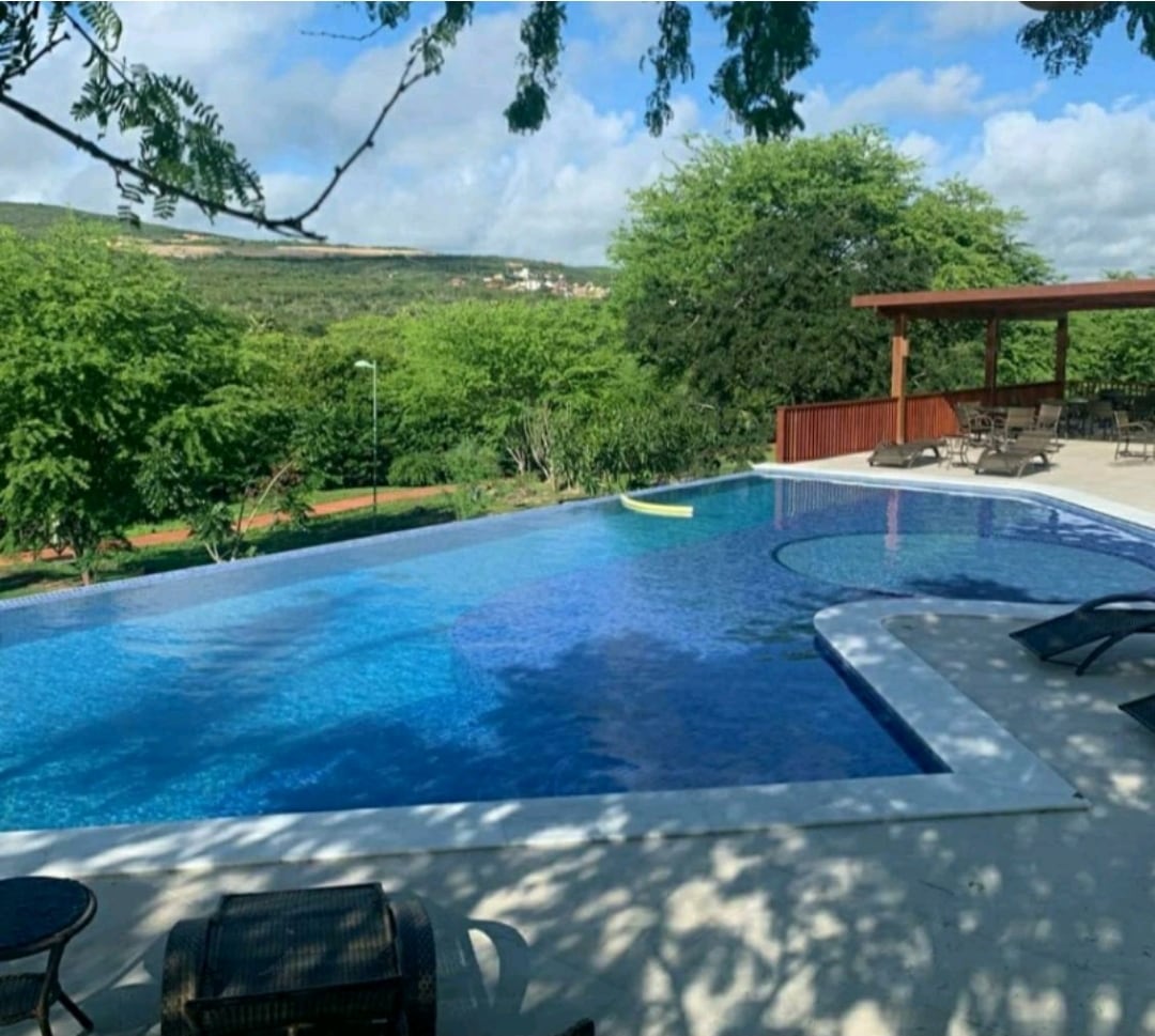 An outdoor pool area features a spacious swimming pool surrounded by greenery. Comfortable lounge chairs are positioned nearby. A shaded structure with seating offers a place to relax and enjoy the view of the landscape beyond.