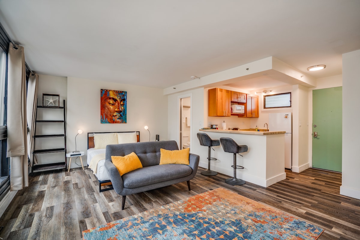 A modern studio features a cozy seating area with a grey sofa and decorative pillows, complemented by a colorful rug. A breakfast bar with two stools overlooks a kitchen with wooden cabinets. Large windows allow natural light to fill the space, enhancing the open layout.