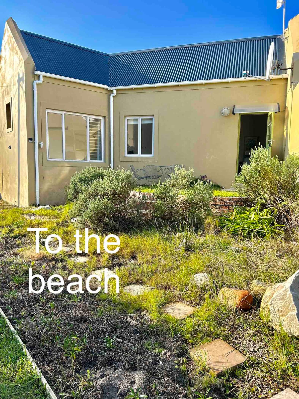 A well-maintained pathway with stepping stones leads towards an entrance. Native vegetation is present in the landscaped area, enhancing the natural setting. The spacious exterior features light-colored walls and a blue roof under clear skies, indicating proximity to the beach.