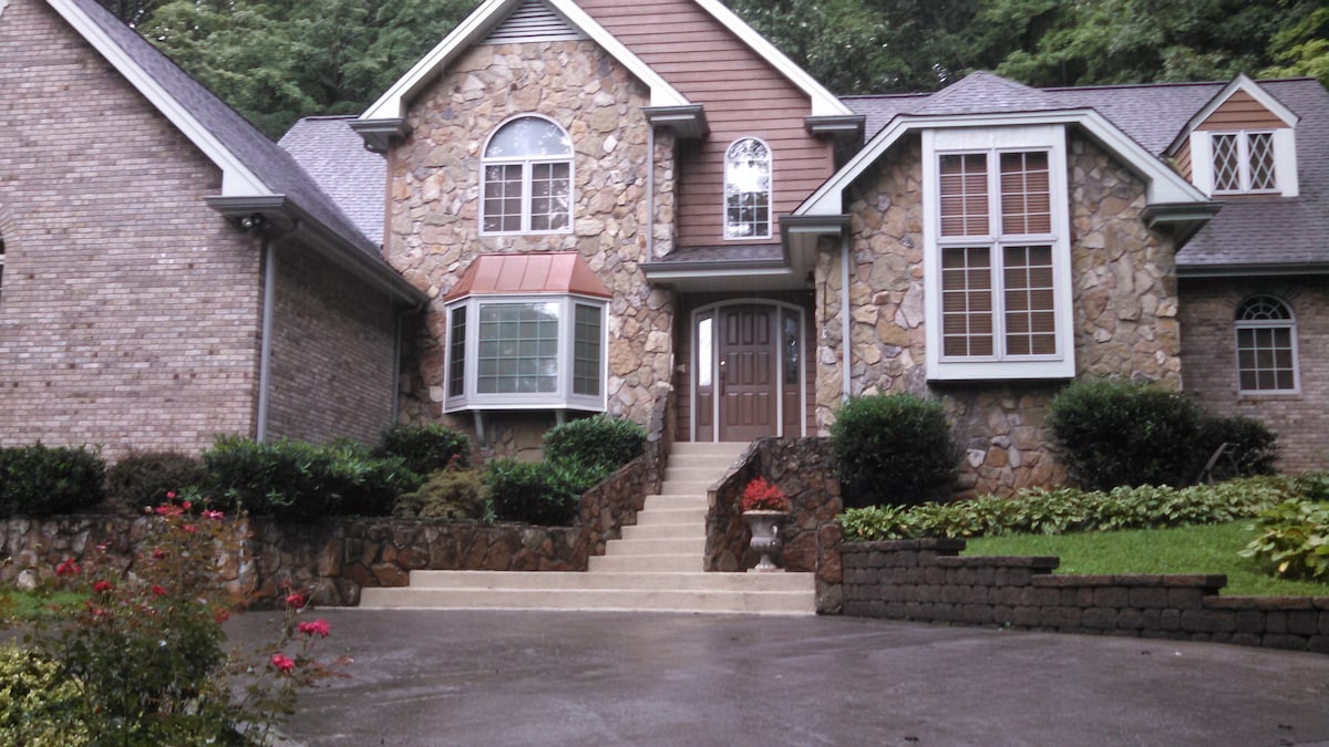 A stone-faced house displays a large, arched front window complemented by traditional rectangular windows. A set of stairs leads to the front door, flanked by manicured greenery and flower beds. The driveway features a smooth surface, providing easy access to the entrance.