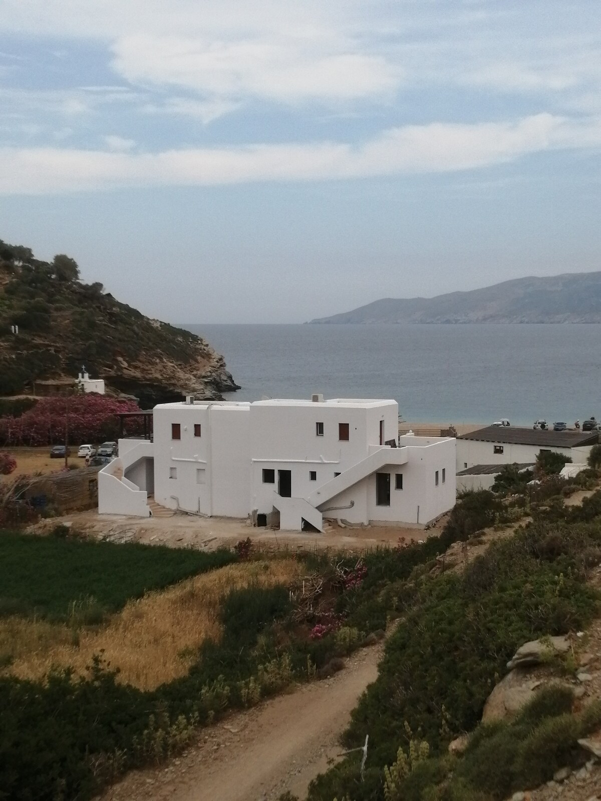A modern white building is positioned near the coastline, featuring unique architectural lines. Surrounding greenery and flowered shrubs provide a natural contrast, while the sea is visible in the background. The landscape includes hills and a distant view of the island’s terrain.