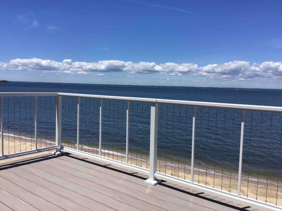 An expansive balcony offers sweeping views of the calm water, with a railing providing safety and unobstructed vistas. The scene is complemented by a partly cloudy sky, capturing the serene ambiance of the beach environment.