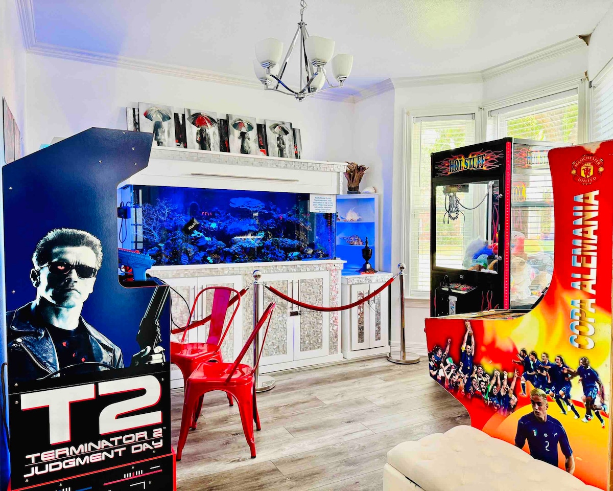 An entertainment area is highlighted by two arcade game machines, with a vibrant saltwater aquarium featuring colorful fish and corals at the center. Red chairs provide seating, and the space is warmly lit by natural light from surrounding windows.