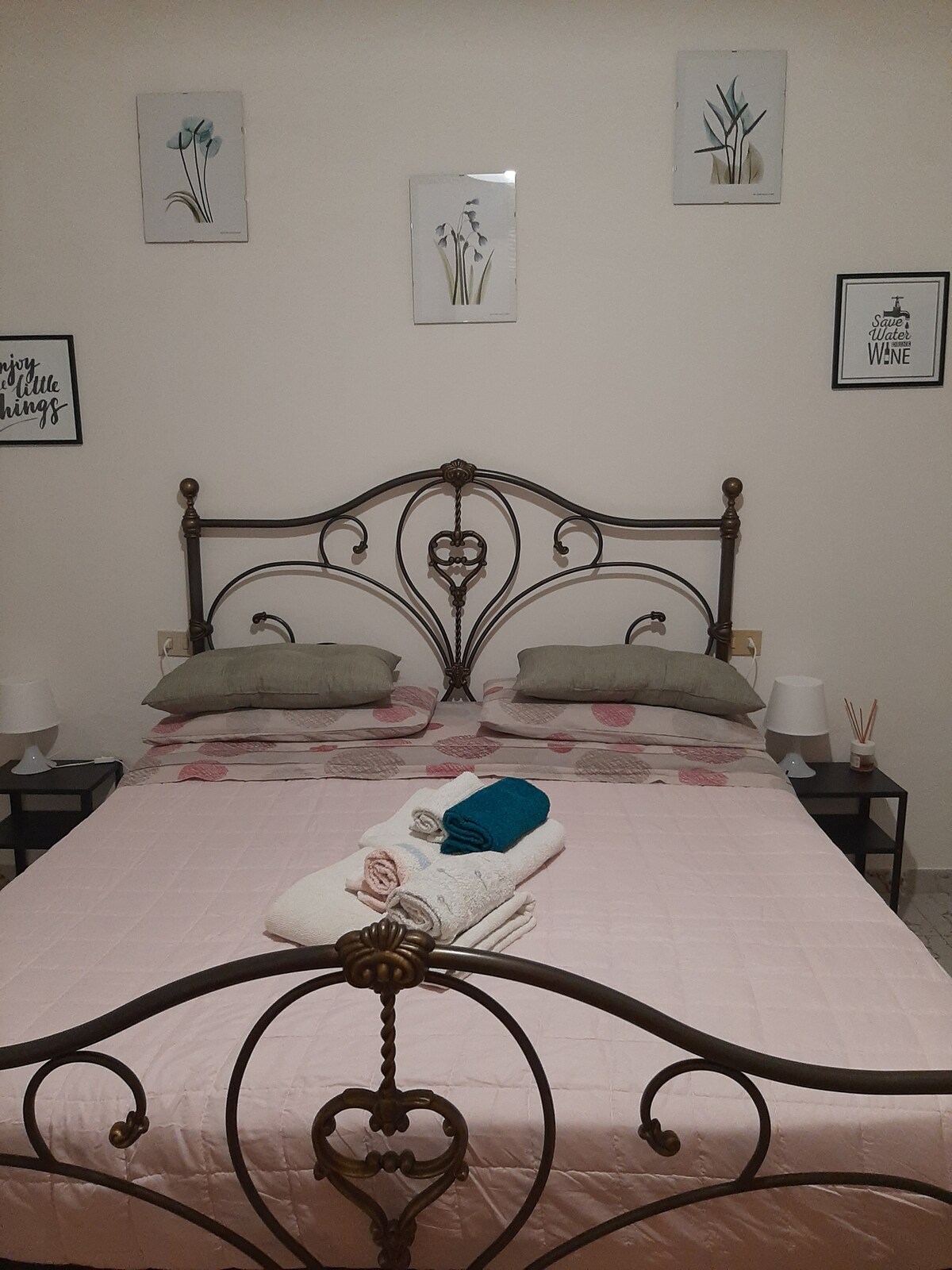 A sleeping area features a decorative metal bed frame with a heart motif. Soft bedding in muted tones is arranged neatly. Two bedside tables with lamps are placed on either side, and wall art depicting floral themes is displayed above the headboard.