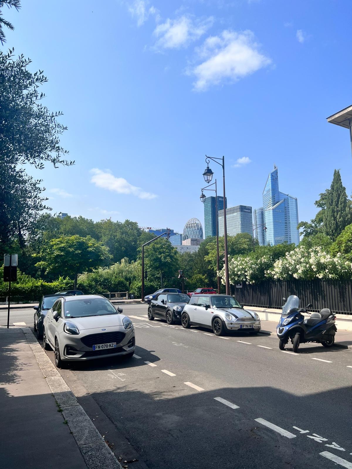 A quiet street scene features a mix of parked vehicles, including a silver car, a black car, and a motorcycle. Lush greenery lines the sidewalk, with a backdrop of modern buildings and a clear blue sky. Street lamps and trees add to the peaceful urban environment.