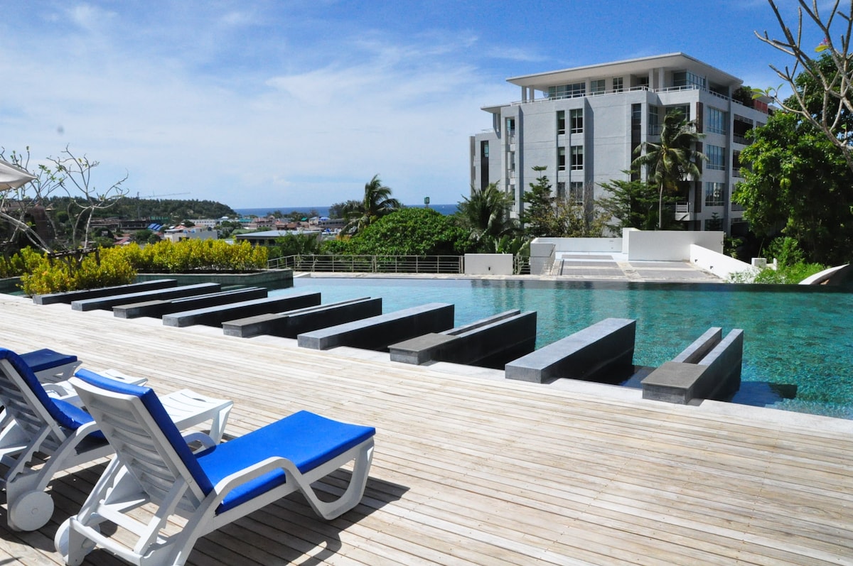 A modern swimming pool is framed by wooden decking, with several lounge chairs positioned beside it. Water features are designed with minimalist elements, and lush greenery is visible in the background, contributing to a serene atmosphere.