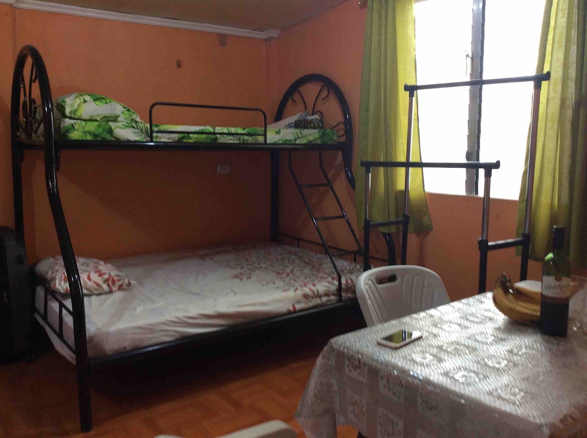 A sleeping area features a black metal bunk bed with a vibrant green and floral-patterned bedding set on the upper level. A spacious bed occupies the lower level, while an adjacent dining table with white plastic chairs provides seating. Large windows are covered by light green curtains.
