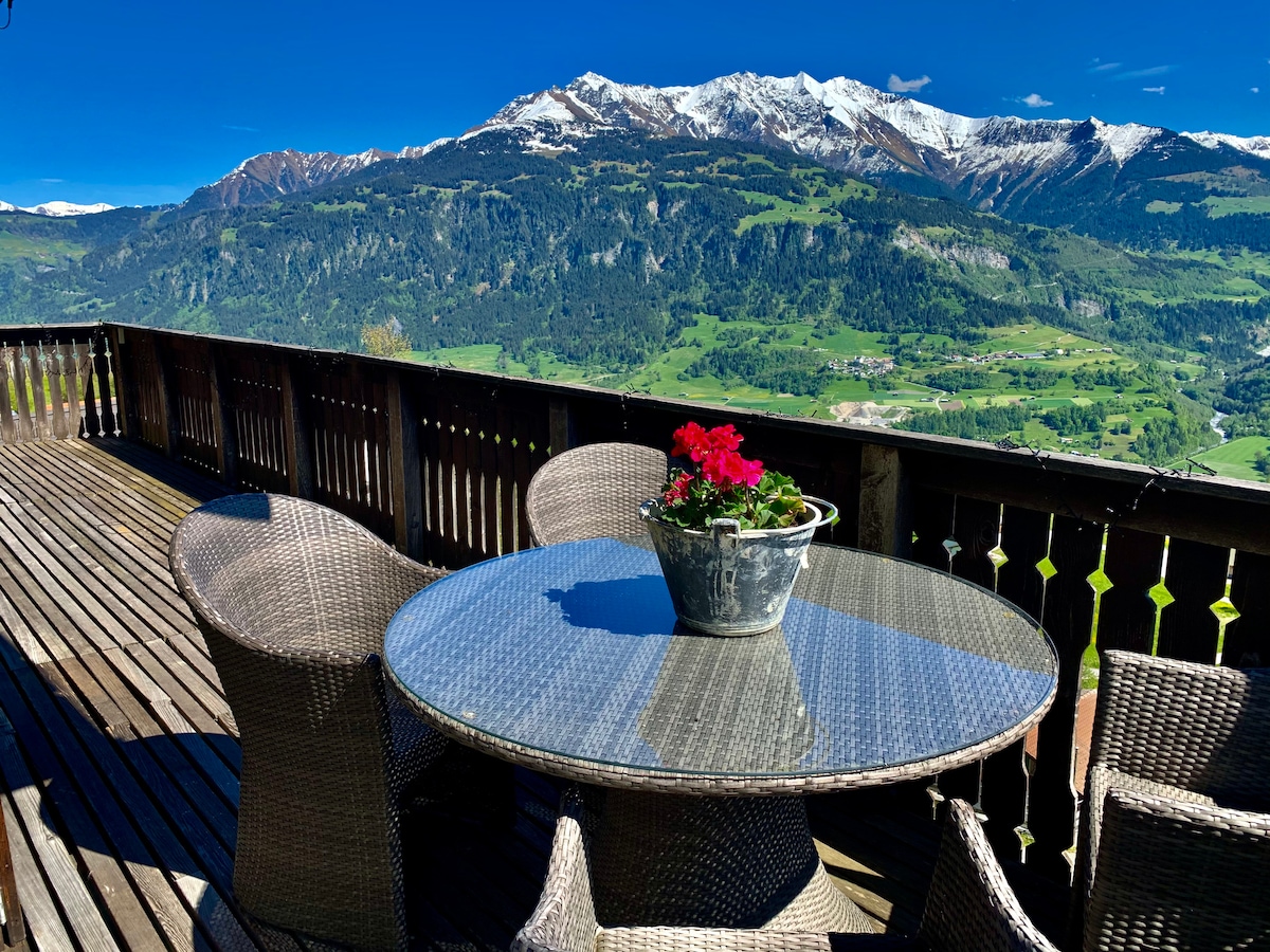 A wooden deck features a round glass-top table surrounded by four woven chairs. A planter with vibrant red flowers is centered on the table. The backdrop showcases a panoramic view of lush green valleys and snow-capped mountains under a clear blue sky.