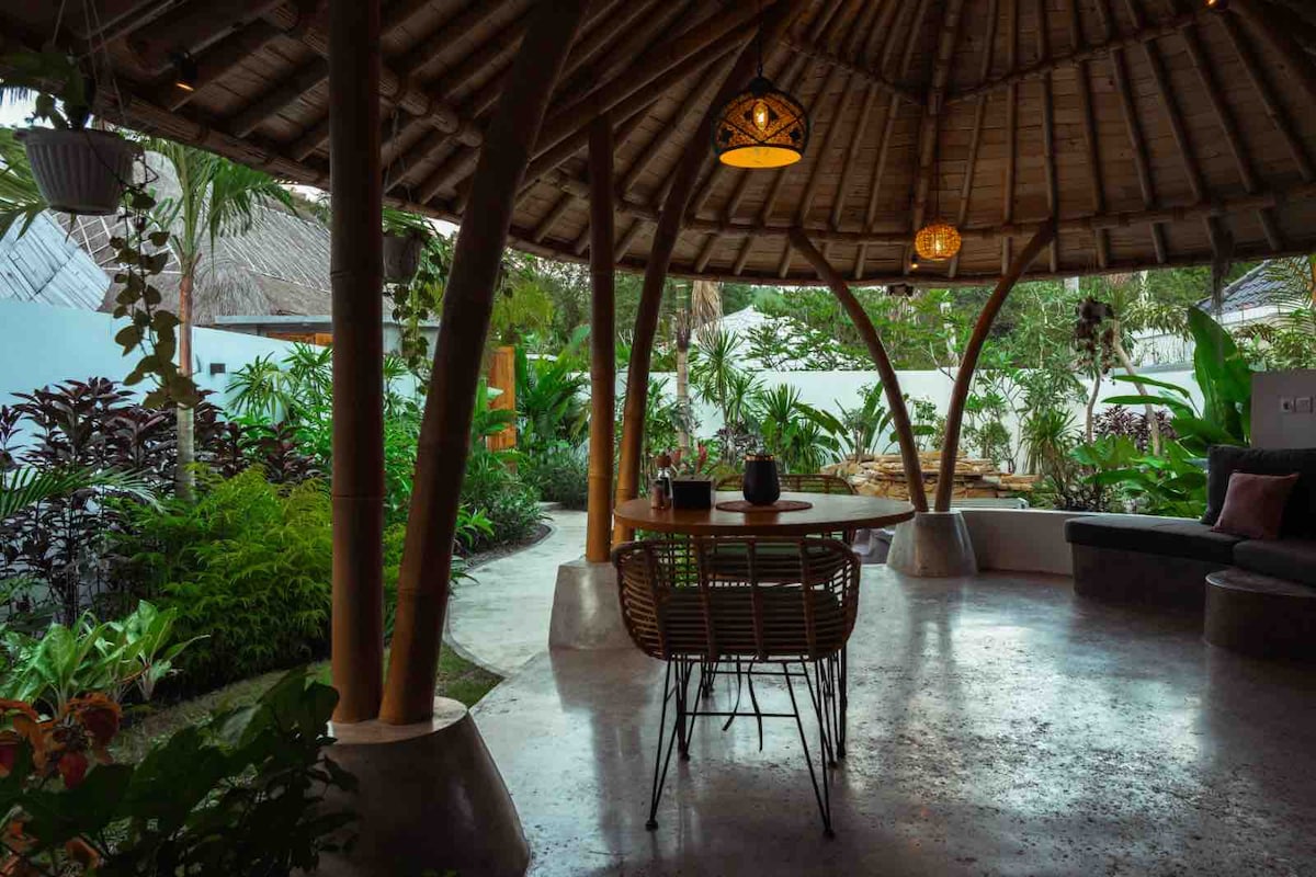 An open-air seating area is set beneath a thatched roof supported by bamboo columns. A round table is surrounded by woven chairs, while lush greenery and tropical plants create a serene ambiance in the background.