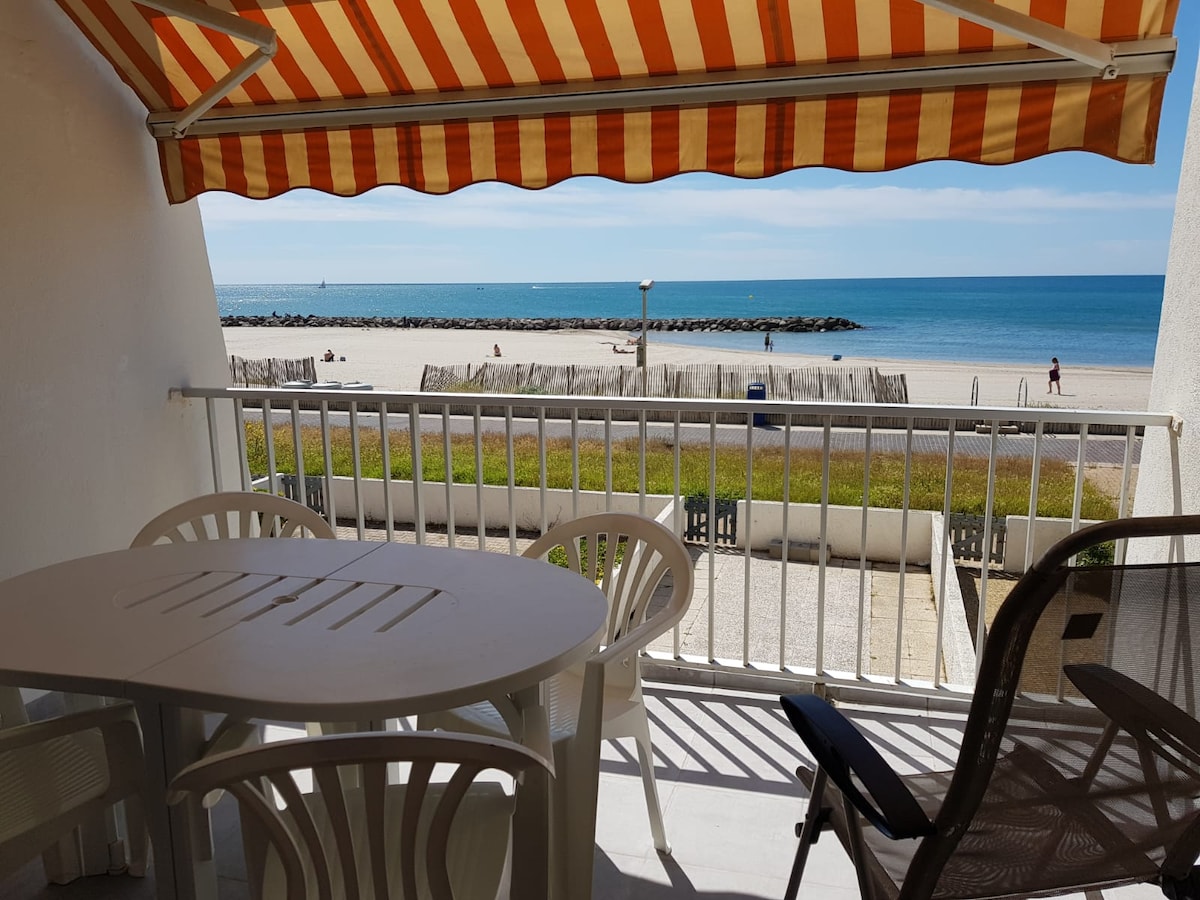 A terrace features a round table and plastic chairs, with a striped awning providing shade. The view extends to a sandy beach and calm waters, with distant silhouettes of beachgoers enjoying the day. A low stone barrier separates the terrace from the coastline.