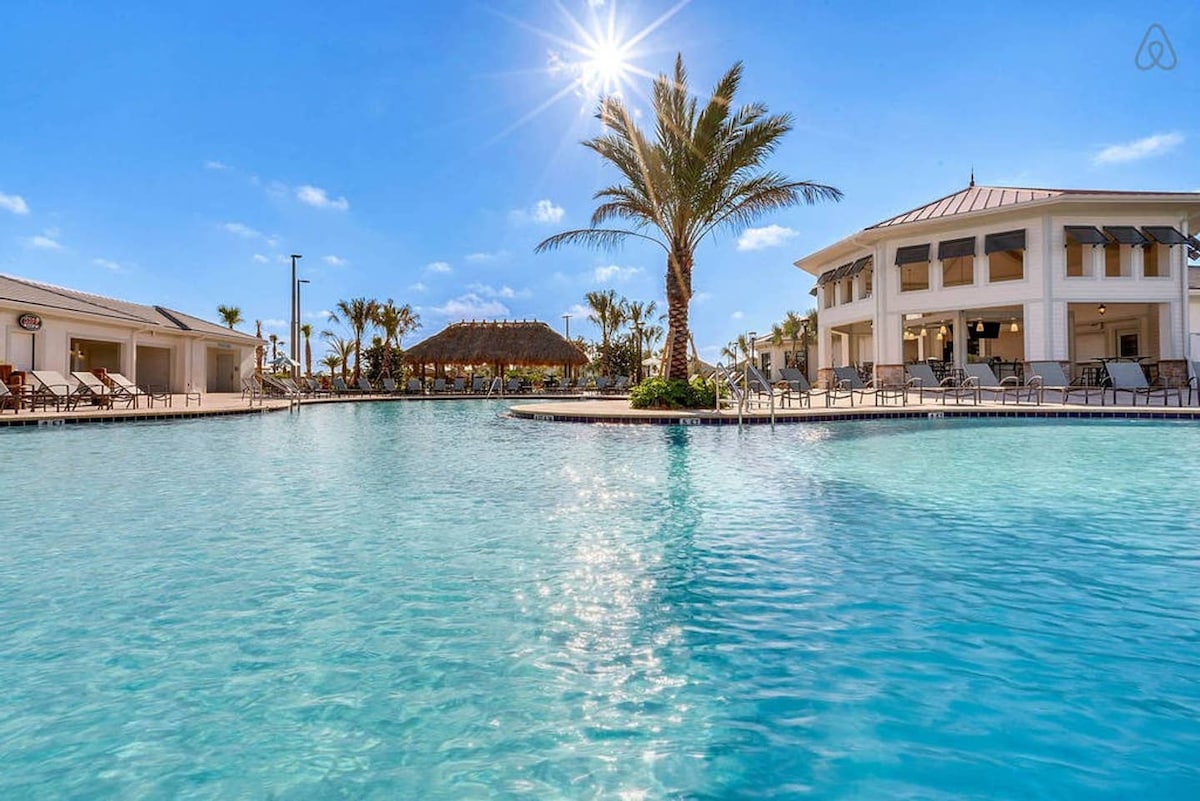A large swimming pool is surrounded by lounge chairs and palm trees, reflecting the sunlight. The clear water invites relaxation, while a clubhouse with a light-colored façade is visible in the background, contributing to the resort-like atmosphere.