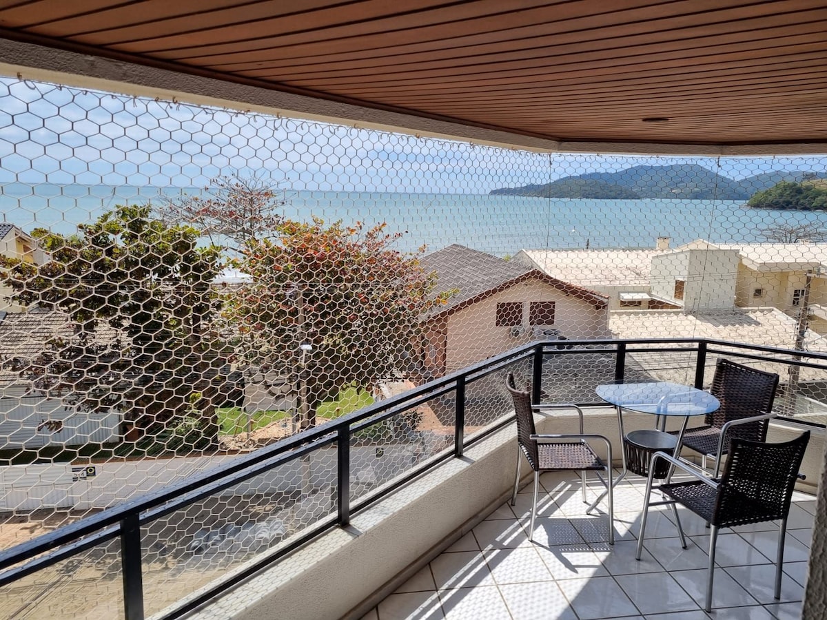 A balcony offers a view of the ocean and nearby trees. A round table is accompanied by four black chairs, providing an inviting space for outdoor relaxation. The balcony is enclosed with a wire mesh for safety, allowing unobstructed views while ensuring security.