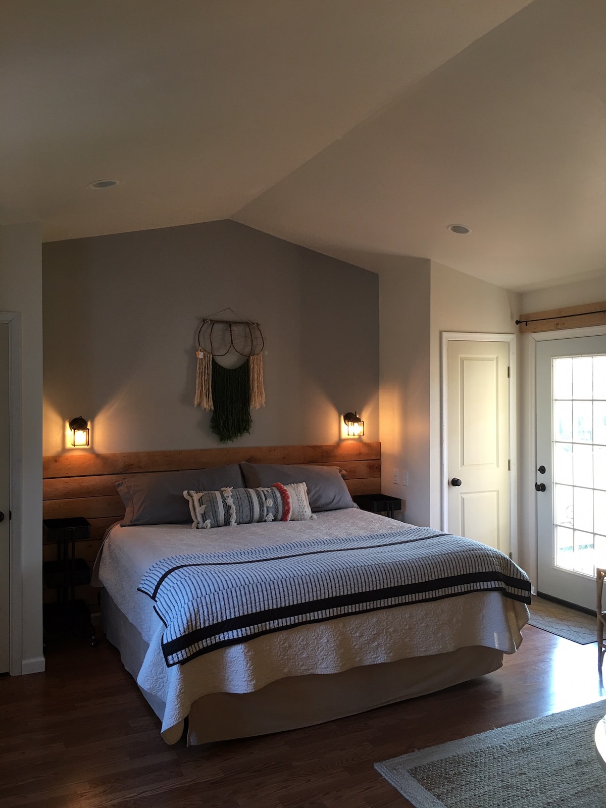 A spacious bedroom features a comfortable bed with decorative pillows, accented by stylish wall sconces. Natural light enters through a glass door leading to the outside. A wooden accent wall offers a warm touch, while simple decor enhances the room's inviting ambiance.