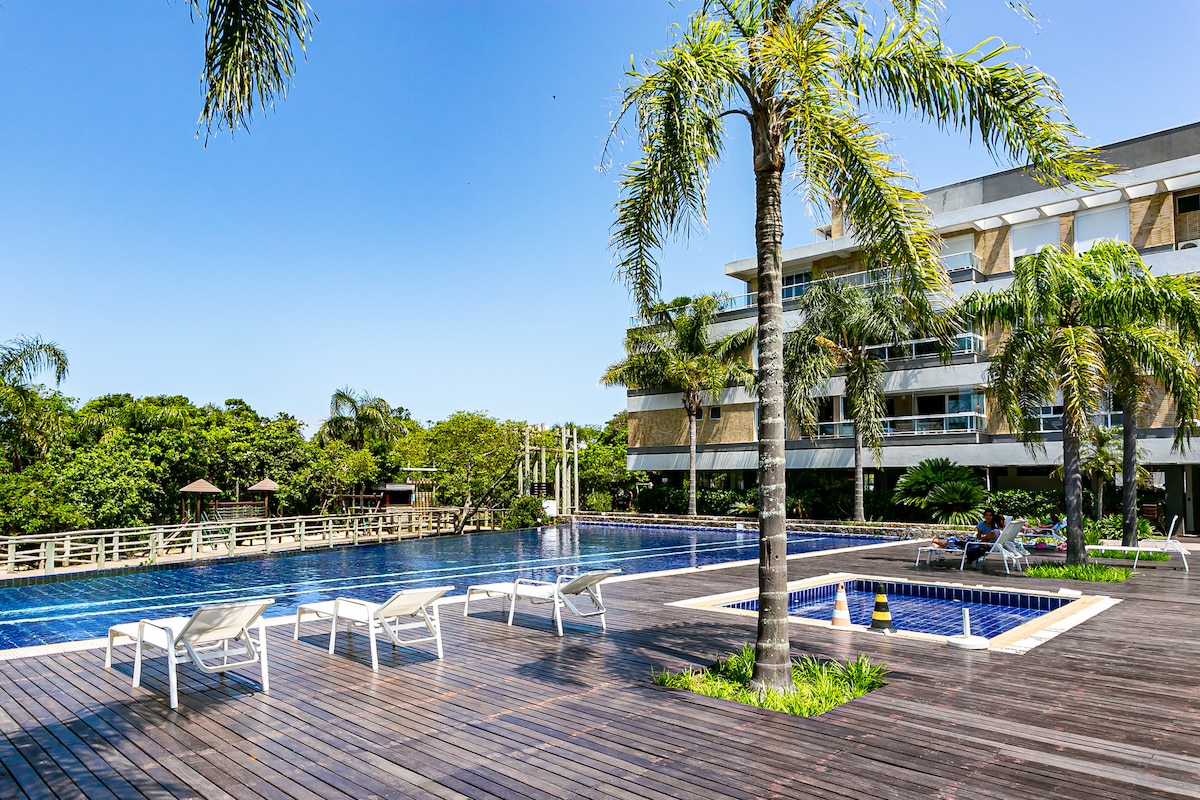 An outdoor swimming pool is surrounded by palm trees and features a wooden deck with lounge chairs. A smaller jacuzzi is adjacent to the pool. Lush greenery is visible in the background, providing a natural setting for relaxation.