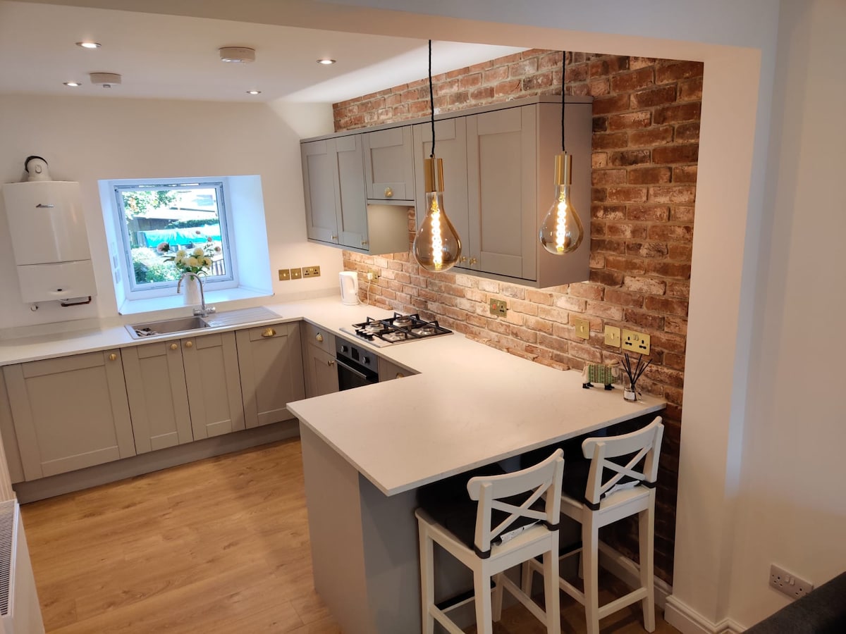 The modern kitchen features light grey cabinetry and a white countertop, complemented by exposed brick walls. Two pendant lights hang above a small breakfast bar with two white stools. A window offers a view of the outside, allowing natural light to illuminate the space.
