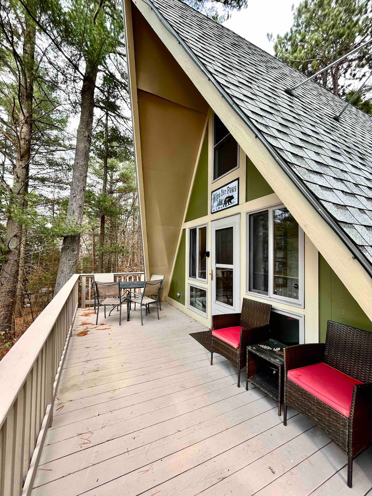 An A-frame chalet is depicted, featuring a spacious front deck furnished with a dining table for four and two small armchairs. The deck overlooks a wooded area, surrounded by tall trees, providing a serene natural setting.