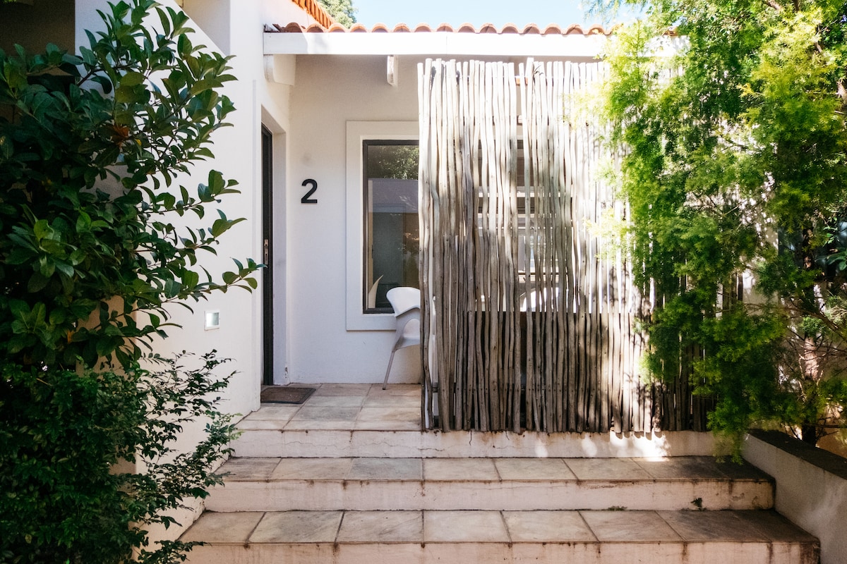 A pathway leads to the entrance of the apartment, flanked by leafy green shrubs. A wooden trellis creates a natural privacy barrier against the seating area, which includes a simple chair. A large window is positioned next to the entrance, allowing light into the space.