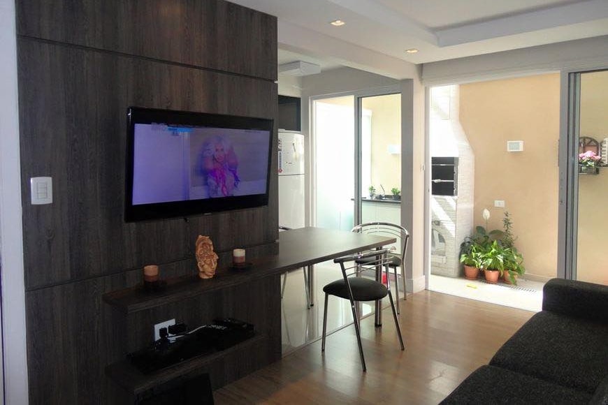 A modern living area features a wall-mounted television opposite a sleek dark wood console. A small dining table with a chair is positioned nearby, and large windows offer a glimpse of a garden area with plants and an outdoor barbecue.
