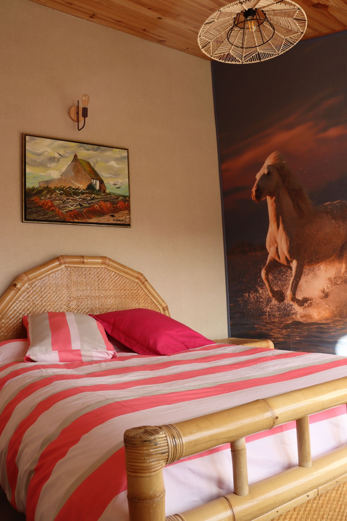 A cozy bedroom features a woven bed frame with a striped pink and white duvet. A large artwork depicting a running horse adorns one wall, while a landscape painting is displayed on another. Natural light is complemented by a rustic overhead light fixture.
