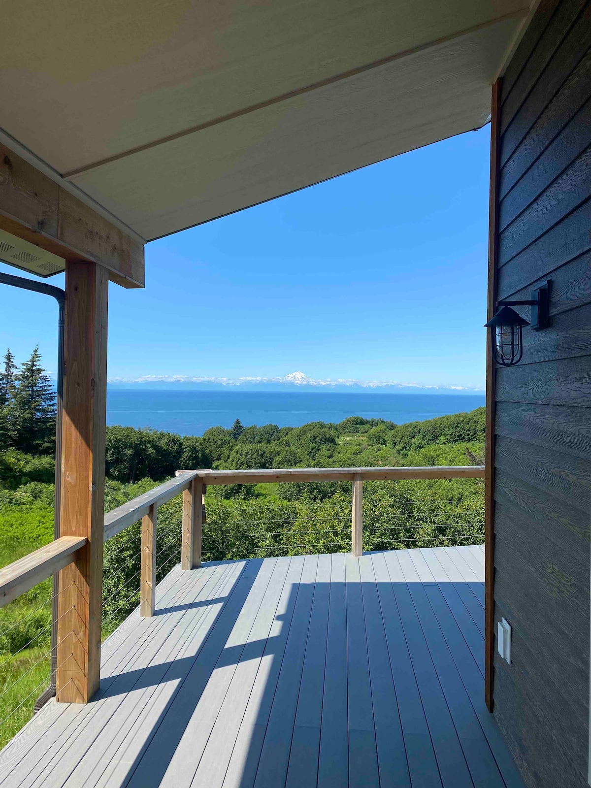 A spacious deck is depicted, offering a clear view of Cook Inlet and distant snow-capped mountains in the background. The sunlit area is surrounded by greenery, enhancing the serene outdoor space. The cabin’s exterior is seen in one corner, with a wall-mounted light fixture.