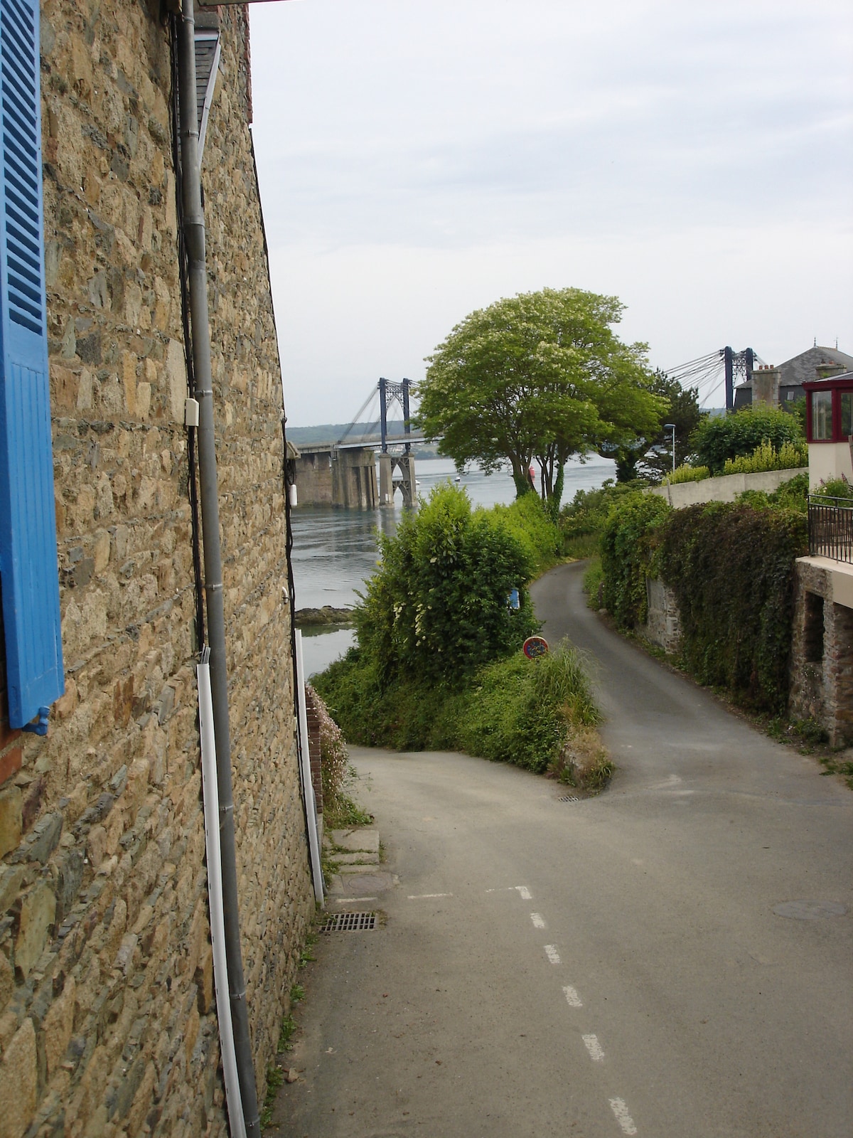 A quiet, winding road leads towards a scenic view of the water, with a bridge visible in the distance. The stone walls of nearby buildings are complemented by lush greenery and trees. The overall setting conveys a peaceful coastal atmosphere.