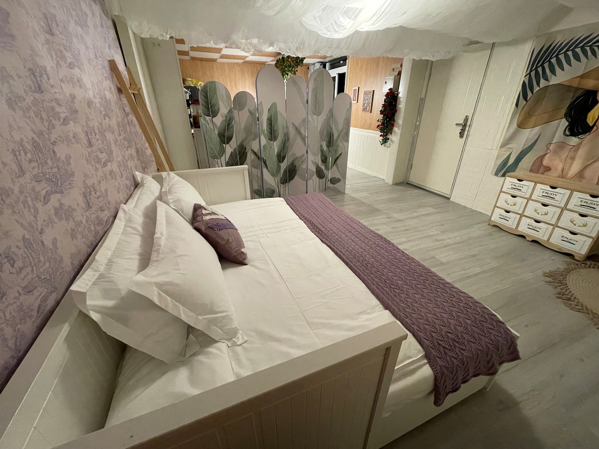 A spacious sleeping area features a bed with crisp white linens and a textured lavender throw. Decorative panels with a botanical design are placed at the foot of the bed, while soft lighting creates a serene ambiance.