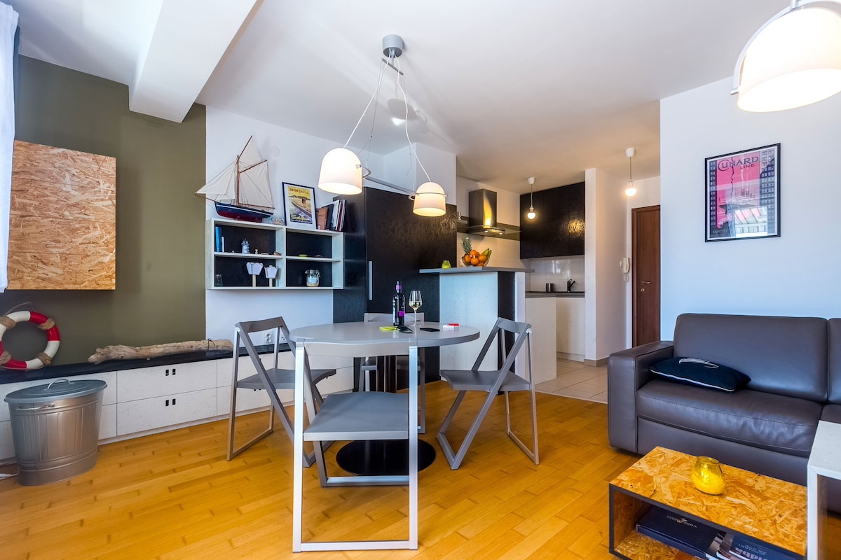 An open living space features a dining area with a round table and grey chairs. A modern kitchen is visible in the background, equipped with counter space and appliances. A leather couch sits nearby, complemented by decorative elements on the shelves, and warm wooden flooring enhances the ambiance.