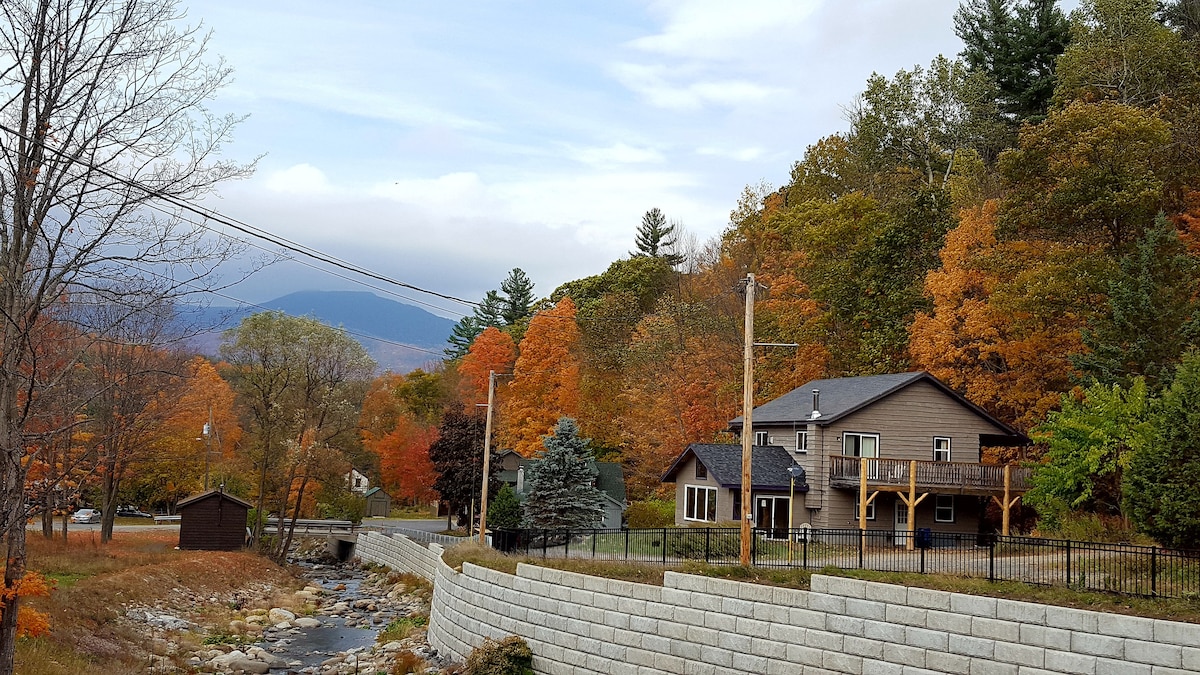 A charming two-story cabin is positioned near a gently flowing stream, surrounded by vibrant autumn foliage. The exterior features a mix of earthy tones and a spacious deck. Mountains can be seen in the background, adding to the natural beauty of the setting.